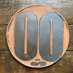 Round, decorative plate with black glaze details and face made from letterpress block impressions on a wooden floor